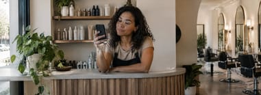 Salon owner at the front desk checking her phone, retail shelves and styling chairs behind her.