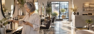 Salon owner counting back bar bottles at a styling station before opening, phone in hand.