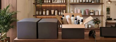  Open shipping box with beauty products on a salon counter, next to unopened boxes and retail shelves in the background.