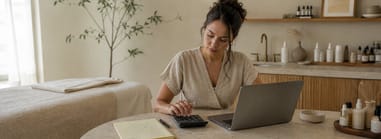 Solo esthetician calculating income and expenses on a laptop and calculator in her treatment room.
