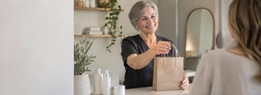 Salon professional handing a retail product bag across the counter to a client at checkout.