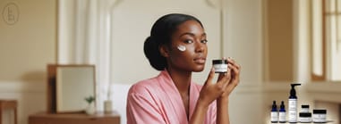 Woman in pink robe applying b.Skincare antioxidant moisturizer, with full product line displayed in foreground.