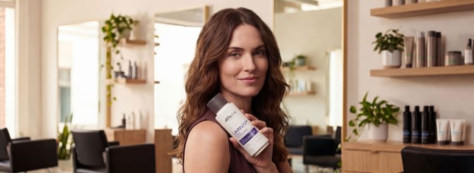 Woman with wavy hair holding Alfheim Lavender+ shampoo bottle in a modern salon interior with plants and shelves in the background.