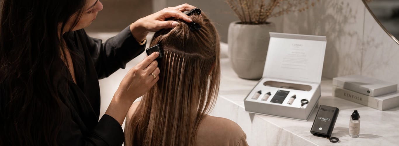 Stylist applying Cosimo Luma UV-bond hair extensions strand by strand, with the kit and curing lamp on the counter.